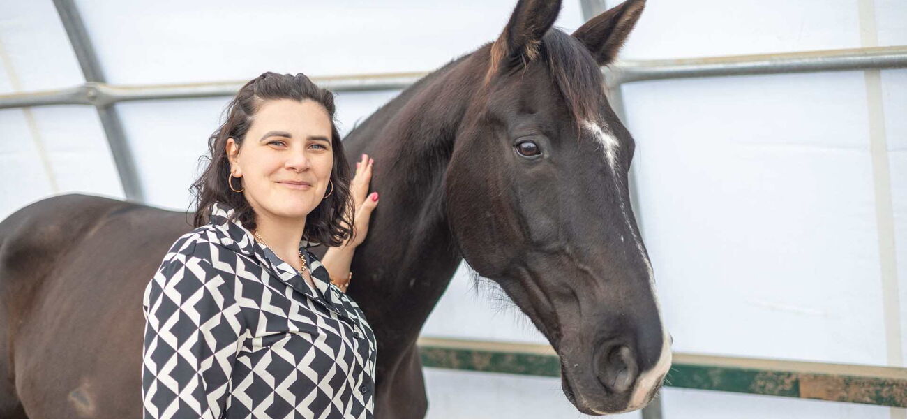 Eine Reitstallbetriebsberaterin steht neben einem schwarzen Pferd mit einer Hand am Hals des Pferdes und lächelt in die Kamera