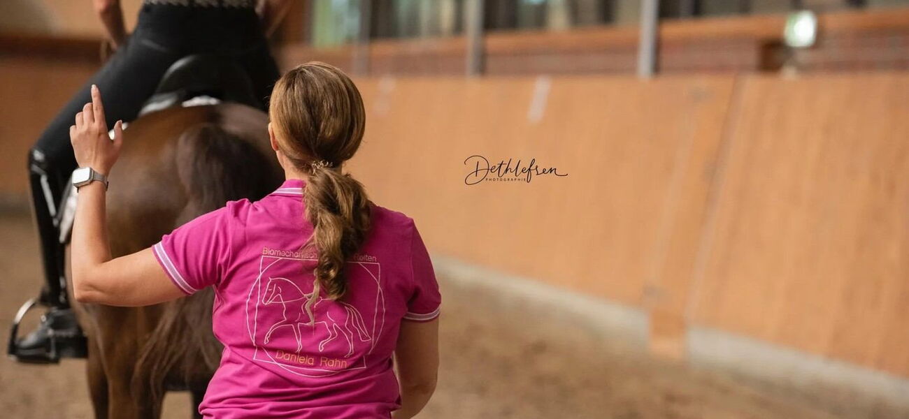 Eine Reitlehrerin betrachtet das Pferd-Reiter-Paar von hinten um den Reitersitz zu beurteilen
