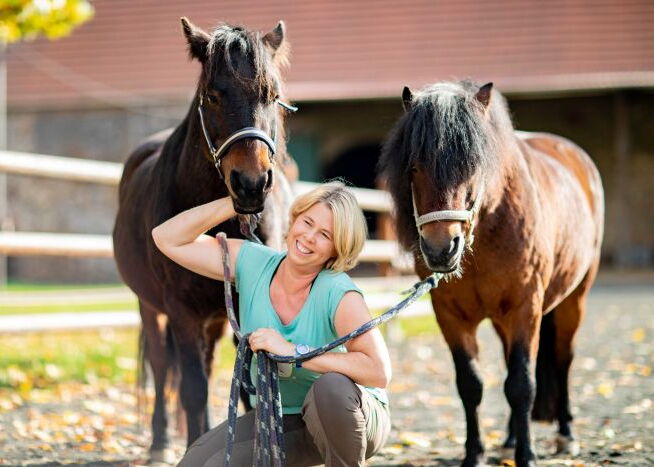 Eine Frau hockt zwischen zwei kleinen Ponys im Sommer und lacht
