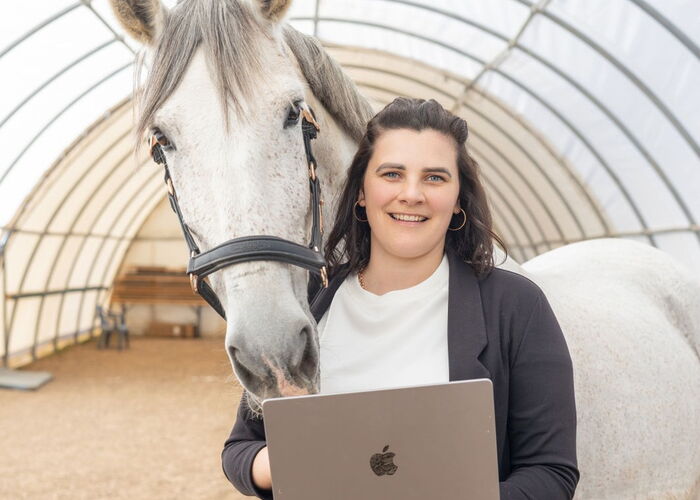 Eine Pferdebetriebsberaterin steht in einer Reithalle mit einem Laptop in der Hand und einem weißen Pferd im Hintergrund
