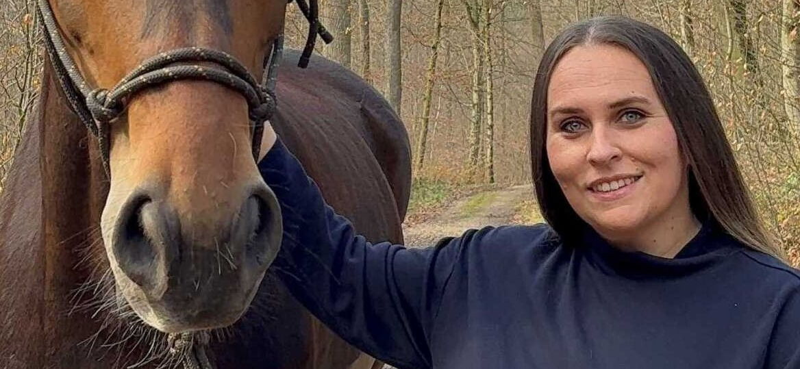 Eine Frau steht neben einem braunen Pferd und lächelt in die Kamera