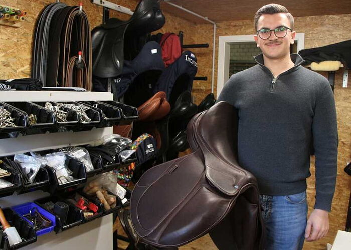 Ein Sattler posiert mit einem Reitsattel auf dem Arm in einem Reitsportgeschäft für ein Foto