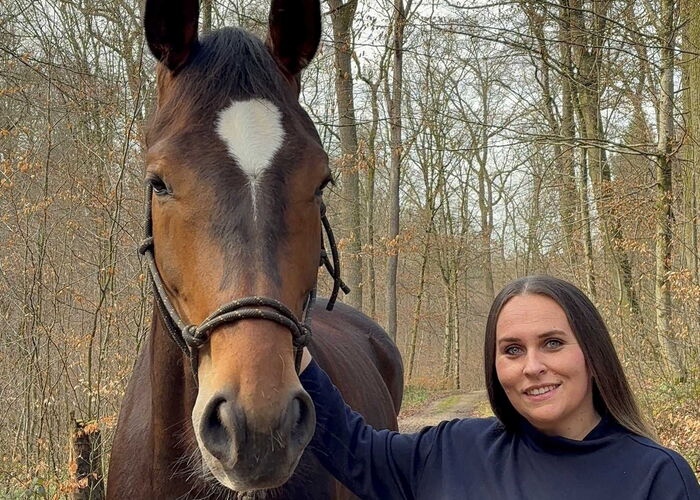Eine Frau steht neben einem braunen Pferd, schaut in die Kamera und streichelt das Pferd am Hals